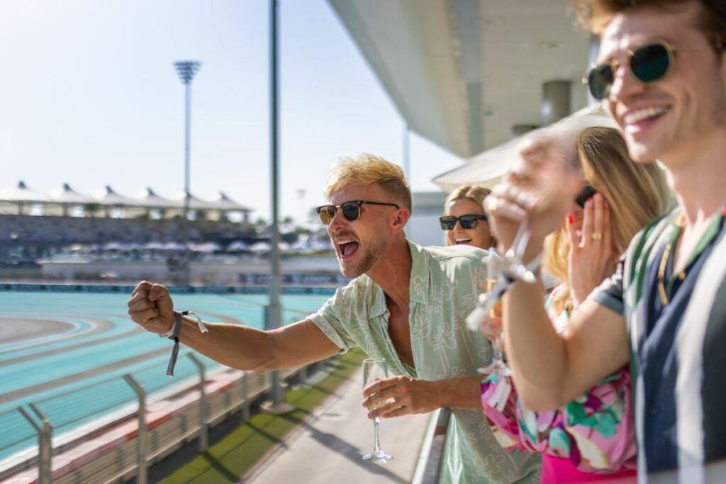 Excited group of people cheering at a race track event, holding drinks and wearing sunglasses.